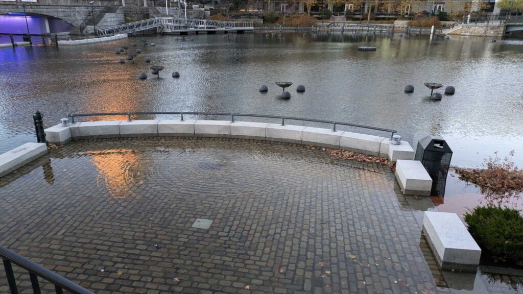 a brick walkway with a brick walkway and a body of water with a bridge and buildings in the (MyCoast AI)